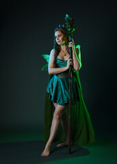 Fototapeta premium Full length portrait of female model wearing fantasy fairy costume, green dress, cloak, butterfly wings and crown. Full length walking pose holding druid staff. isolated on dark studio background.