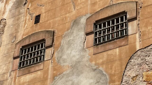 An old, dilapidated building with barred windows. The rough facade and metal bars create a strong atmosphere of constraint, isolation, and control. A prison, an abandoned institution, or a historical