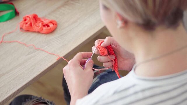 Close-up of woman's hands crocheting doll clothes using a hook and yarn. Creative handmade craft hobby and DIY activity while watching online lessons on a laptop at home. 4K POV shot