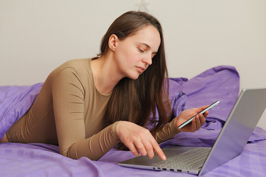 A woman lies on a bed with a purple blanket. She types on a laptop with one hand and holds a smartphone with the other hand while focused on her task