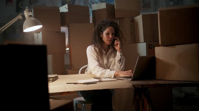 Female small business owner talking on mobile phone while working on laptop in warehouse office.