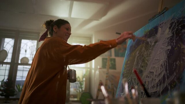 Smiling young female artist painting on large canvas in her bright creative studio.