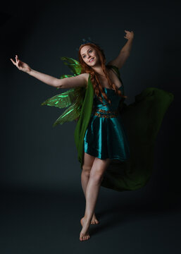 portrait of female model wearing fantasy fairy costume, green dress, cloak, butterfly wings and crown. full length standing pose, isolated on dark studio background.