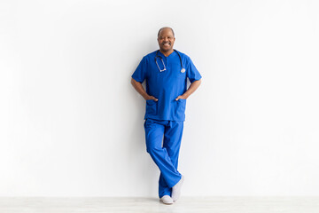 Obraz premium Confident mature African American male doctor in blue scrubs and stethoscope leaning casually against white wall. Full body professional medical portrait on studio background