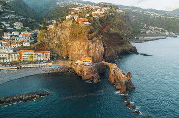 Aerial view of Ponta do Sol village in Madeira, Portugal, showing colorful buildings on a rocky cliff overlooking the Atlantic Ocean and a stone pier structure. © JULIA