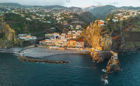An aerial shot captures the coastal village of Ponta do Sol in Madeira, featuring colorful buildings nestled between steep cliffs and a pebble beach by the Atlantic Ocean.