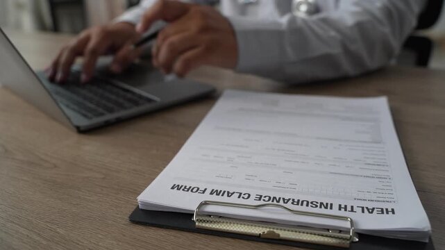 Doctor hand holds pen on health insurance claim form with stethoscope on clipboard, explaining medical policy details and healthcare coverage to patient on wooden office desk.