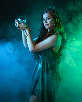 close up portrait of female model wearing fantasy fairy costume, green dress, butterfly wings and crown. standing pose holding  magical glass orb.  glowing green  lighting, dark studio background