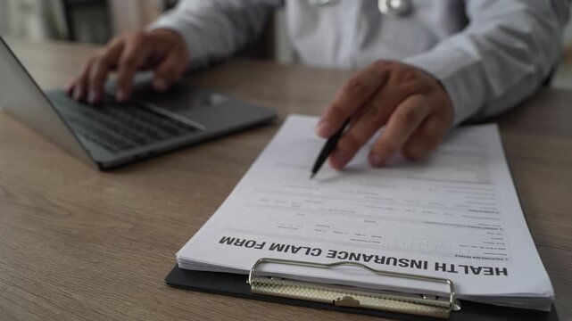 Doctor hand holds pen on health insurance claim form with stethoscope on clipboard, explaining medical policy details and healthcare coverage to patient on wooden office desk.
