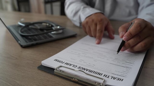 Doctor hand holds pen on health insurance claim form with stethoscope on clipboard, explaining medical policy details and healthcare coverage to patient on wooden office desk.