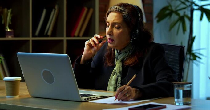 Senior Indian businesswoman in corporate attire taking notes while attending training session on laptop in modern cozy office space experienced female executive using headphones with mic at desk