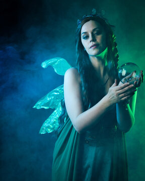 close up portrait of female model wearing fantasy fairy costume, green dress, butterfly wings and crown. standing pose holding  magical glass orb.  glowing green  lighting, dark studio background