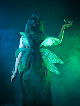 close up portrait of female model wearing fantasy fairy costume, green dress, butterfly wings and crown. standing pose holding  magical glass orb.  glowing green  lighting, dark studio background