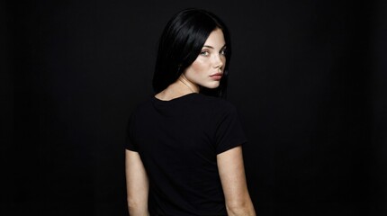Fototapeta premium A young Caucasian woman with long black hair looks over her shoulder against a dark background. She wears a simple black t-shirt and has a serious expression.