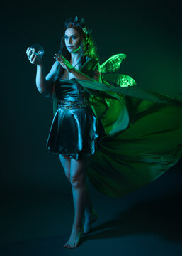 portrait of female model wearing fantasy fairy costume, green dress, cloak, butterfly wings and crown. full length standing pose, holding  magical crystal orb.  isolated on dark studio background.