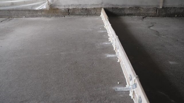Static slow pan shot of a construction site. A wooden board serves as formwork a screed guide on a rough concrete floor, secured with metal brackets expanding foam. Raw industrial background