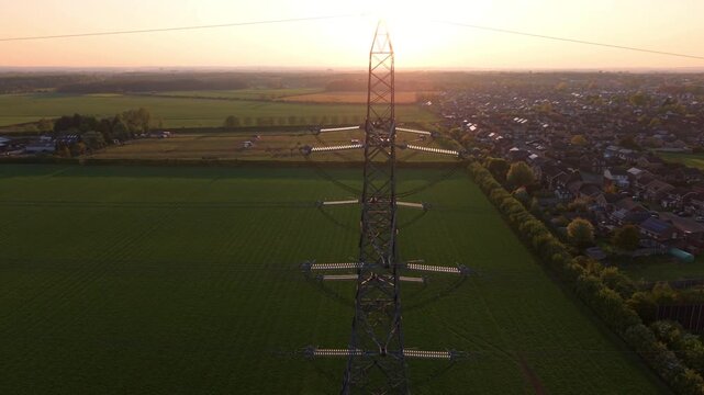 High angle aerial drone view close up of a steel lattice high voltage electricity pylon overlooking rural agricultural fields during a golden hour sunset near Doncaster UK