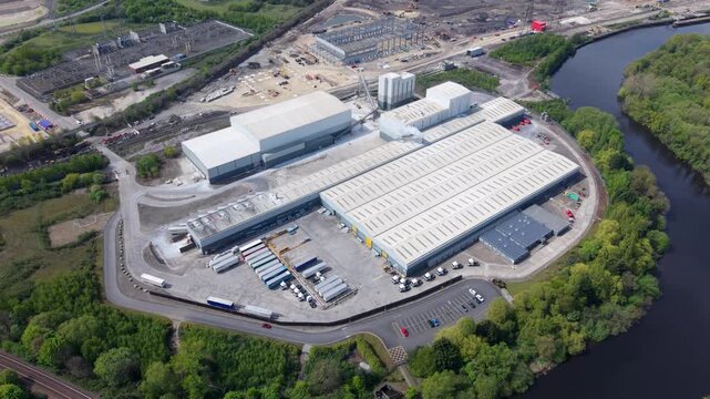 Industrial manufacturing warehouses and logistics yard with commercial HGVs and smoke from an exhaust stack, aerial view, UK.
