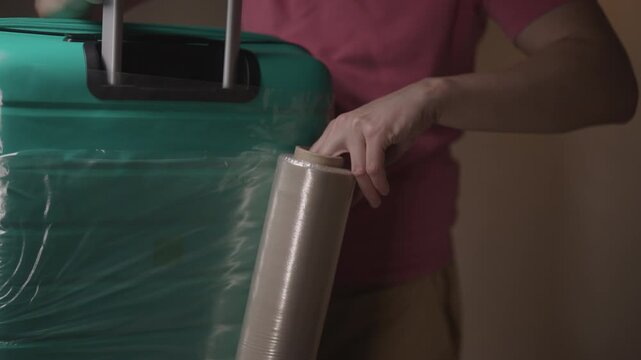 Traveler preparing for vacation by wrapping turquoise suitcase with plastic shrink wrap to protect baggage during flight and prevent theft. Concept of travel and packing for upcoming holiday.