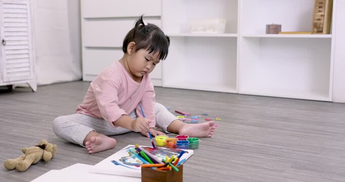 Asian preschool daughter painting independently with colorful brush in slowmotion while sitting barefoot on floor developing focus, imagination and motor skills through early childhood art play
