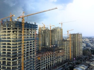Fototapeta premium High-rise building rises against a stormy sky. Cranes loom over active construction sites. Urban development pulses with progress and ambition. Sky's dark clouds hint at weather's unpredictability