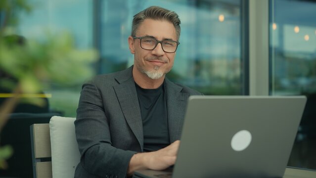 Mature professional man in business casual smiling, working on laptop in contemporary office. Portrait of happy mid adult man with approachable friendly smile. Successful senior entrepreneur.
