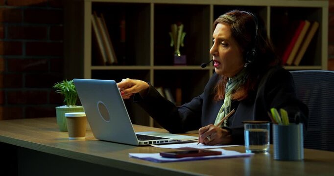Senior Indian businesswoman in corporate attire taking notes while attending training session on laptop in modern cozy office space experienced female executive using headphones with mic at desk