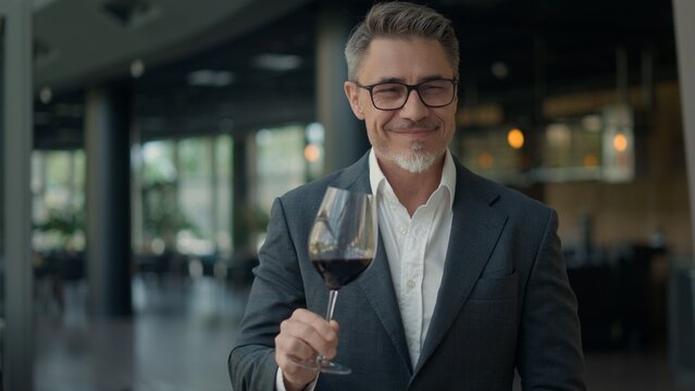 Businessman in suit sitting in a modern restaurant, drinking, tasting red wine. Elegant man holding glass, focusing on evaluating, rating wine. Moment of pleasure and well deserved relaxing break.