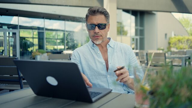 Mature businessman relaxing on cafe terrace, smoking a cigar working on laptop. Mid adult man in sunglasses outdoor. Freedom, connectivity in contemporary business lifestyle. Successful entrepreneur.