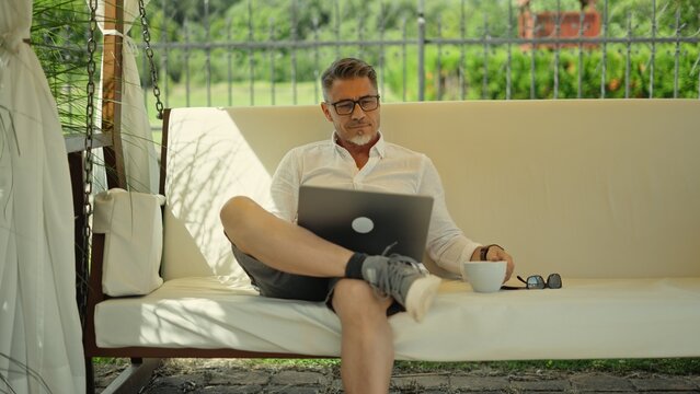 Businessman working on laptop computer, sitting on swinging bench in garden at home. Busy male entrepreneur on summer vacation. Casual mid adult man relaxing, enjoying free business lifestyle.