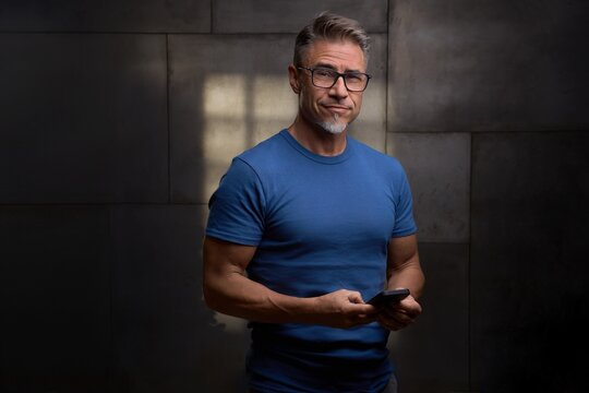 Confident mature man standing against dark industrial wall in office, using mobile phone. Portrait of mid adult handsome male in glasses, smiling. Staying fit and sporty in older age.