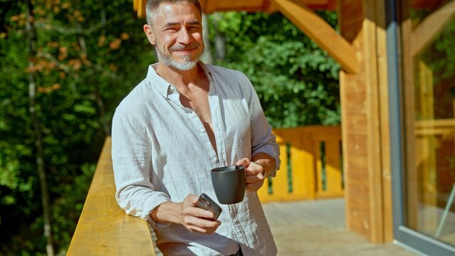 Businessman starting the day with morning coffee and checking phone. Portrait of middle aged man with gray hair in casual cloth outdoor on home balcony. Spring or summer. Happy satisfied smile.
