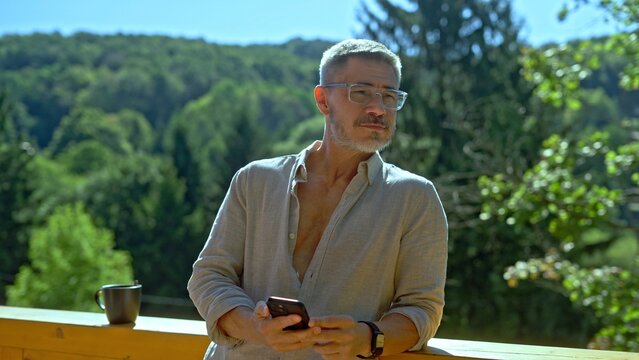 Businessman checking phone reading business news and email at morning. Portrait of middle aged man in eyeglasses and casual cloth outdoor on home balcony. Spring or summer.