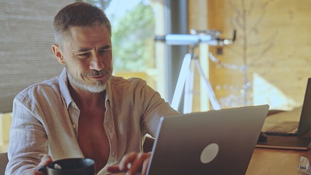 Happy businessman remote working on laptop online in home office, thinking, smiling. Casual middle aged man sitting at desk in living room. Spring or summer. Entrepreneur on getaway in wooden cabin.