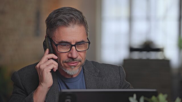 Serious businessman working in home office talking on phone call. Middle aged, older male in glasses concentrating, using laptop and mobile. Telecommunication, entrepreneurship.