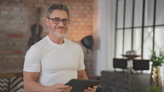 Middle aged man using digital tablet at home in loft living room. Happy, sporty, active senior male with gray hair and glasses reading social media, working and smiling.