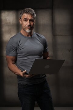Casual mid adult man standing holding laptop computer. Portrait of middle aged male entrepreneur in 50s with gray hair. Businessman in business casual on gray background.