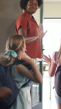 Vertical video: Greeting teacher guiding kids into classroom as girl nears, backpacks and rust blo