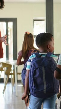Vertical video: Walking kids following teacher in rust top gesturing into classroom with backpacks