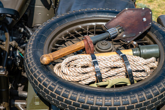 Close-up of a spare wheel on a vintage military motorcycle featuring an attached entrenching shovel, stick grenade, and coiled rope, all secured with leather straps against an olive drab frame.