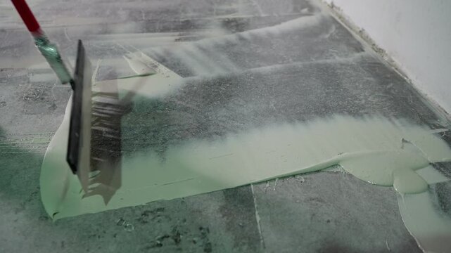 Close up shot of worker spreading pale green epoxy resin or self leveling compound on concrete floor with squeegee. Industrial flooring application and renovation process in progress