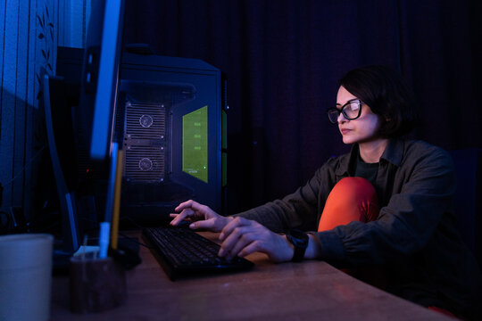 Young woman focused on her computer screen in a dark room with blue ambient light, engaged in a digital project.