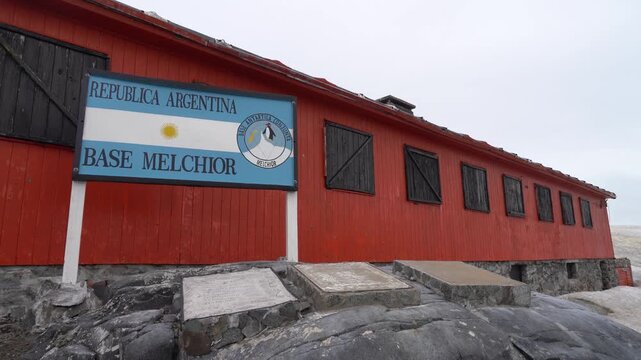 A wide shot of the iconic orange wooden main building at Melchior Base, featuring the official Republic of Argentina sign and commemorative plaques on the rocky ground