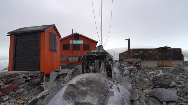 Stationary shot of the orange wooden huts and the Argentine flag at Melchior Base on Gamma Island, set against the rocky Antarctic landscape