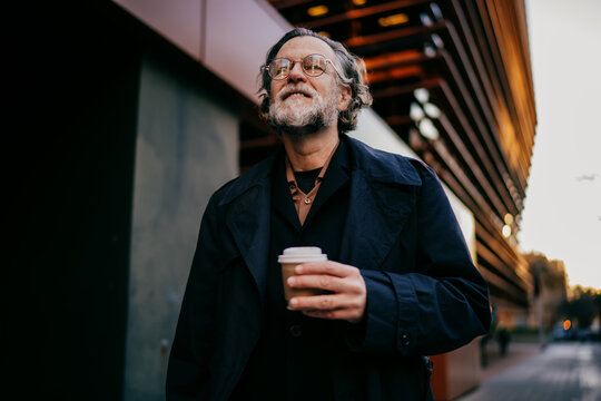 Happy middle aged man with gray hair enjoying walk in evening city holding coffee to go, low angle view of smiling successful senior businessman in glasses and stylish black coat outdoors, copy space.
