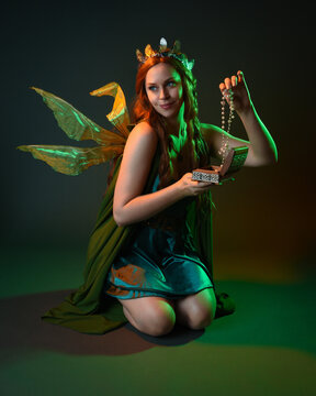 portrait of female model wearing fantasy fairy costume, green dress, cloak, butterfly wings and crown. Full length kneeling pose holding secret jewellery box, isolated on dark studio background