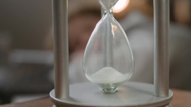Close-up tilt-down shot of sand falling down in hourglass while unrecognizable woman napping in background
