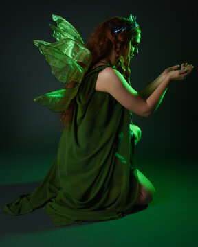 portrait of female model wearing fantasy fairy costume, green dress, cloak, butterfly wings and crown. full length sitting pose, kneeling on floor, dark studio background.