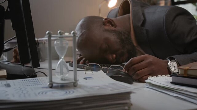 Handheld zoom in shot of African American man sleeping at his office computer desk while tracking time with hourglass