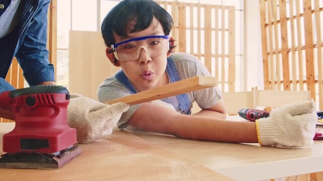 A child wearing goggles and gloves sands and blow a wooden board with an electric sander in a bright workshop, focusing on a DIY carpentry project amid wood framing and sawdust.